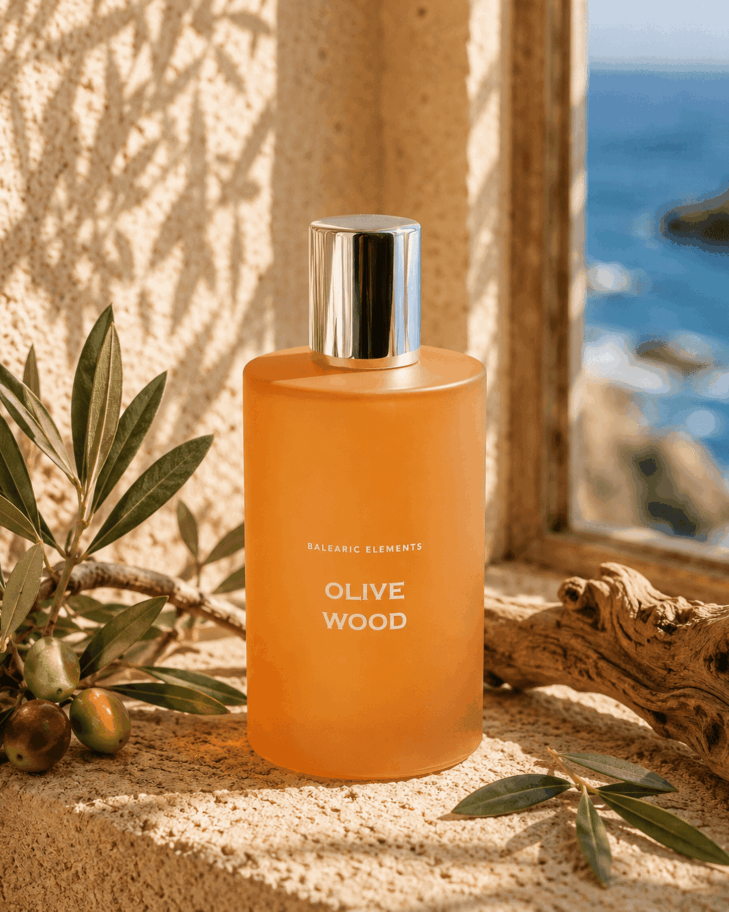 A frosted amber-orange bottle labeled 'BALEARIC ELEMENTS OLIVE WOOD' with a reflective silver cap, set on a sunlit stone ledge with olive branches and driftwood, with the ocean visible in the background.