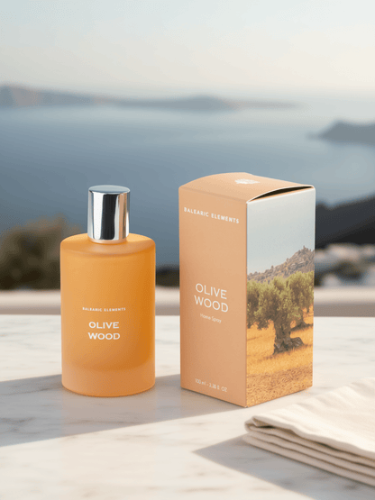 Olive Wood room spray fragrance bottle and packaging box by Balearic Elements, set on a light stone surface with a blurred coastal landscape in the background.