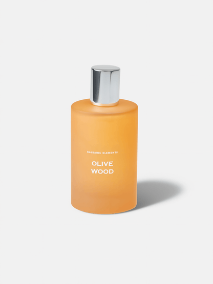 An orange frosted glass bottle with a silver metallic cap, labeled 'OLIVE WOOD' and 'BALEARIC ELEMENTS', on a light grey background.