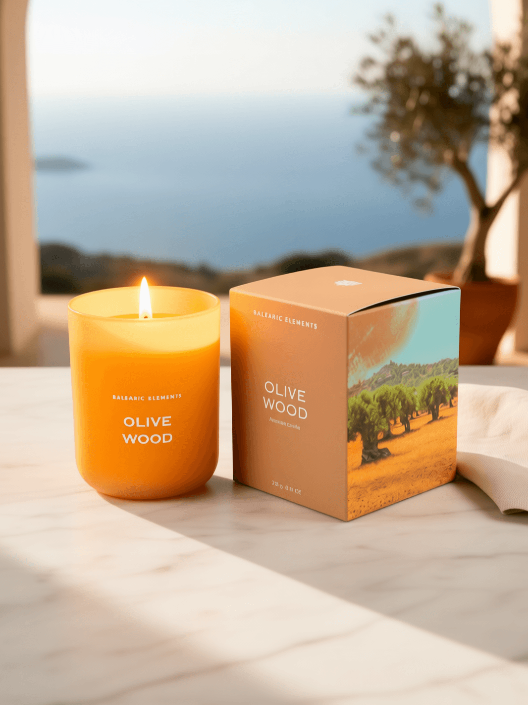 A lit candle in a clear glass jar sits beside its branded box on a marble surface, with a blurred coastal scene and tree in the background. The candle reads 'OLIVE WOOD' 