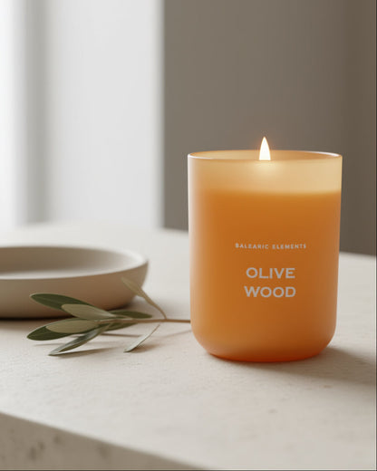 Scented candle labeled 'Olive Wood' on a surface with a bowl and olive branch in the background