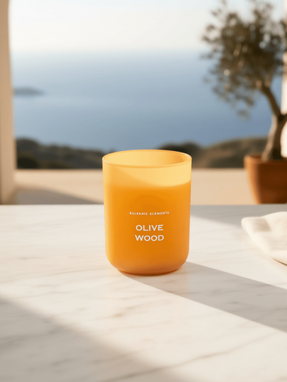 An amber glass candle jar labeled 'OLIVE WOOD' sits on a light marble surface with a blurred coastal terrace background featuring an archway, sea, and potted tree.