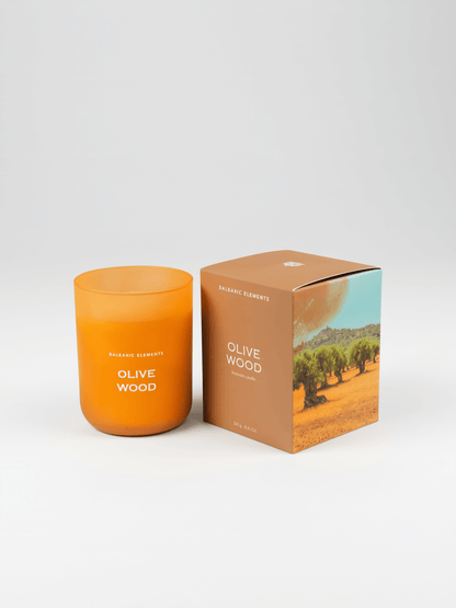 An orange glass jar candle labeled 'OLIVE WOOD' stands next to its brown cardboard packaging box featuring an olive tree landscape print, on a white studio background.