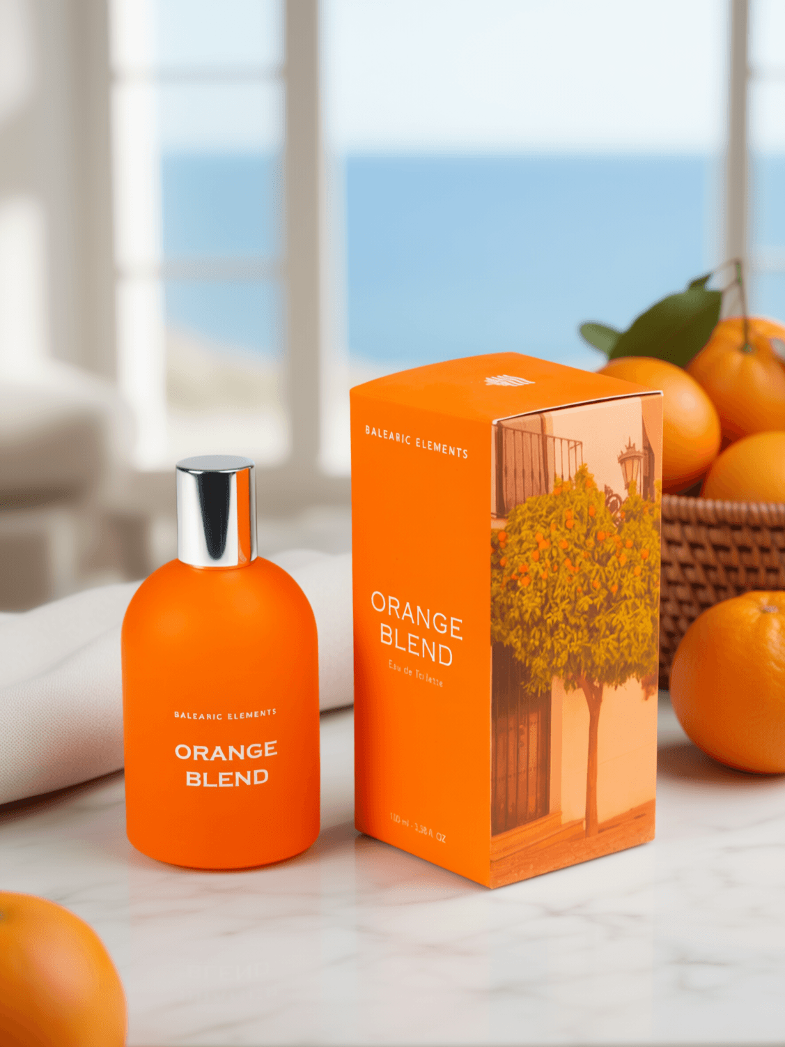 An orange perfume bottle labeled 'BALEARIC ELEMENTS ORANGE BLEND' stands next to its matching orange box on a marble surface, with oranges and a blurred window background.