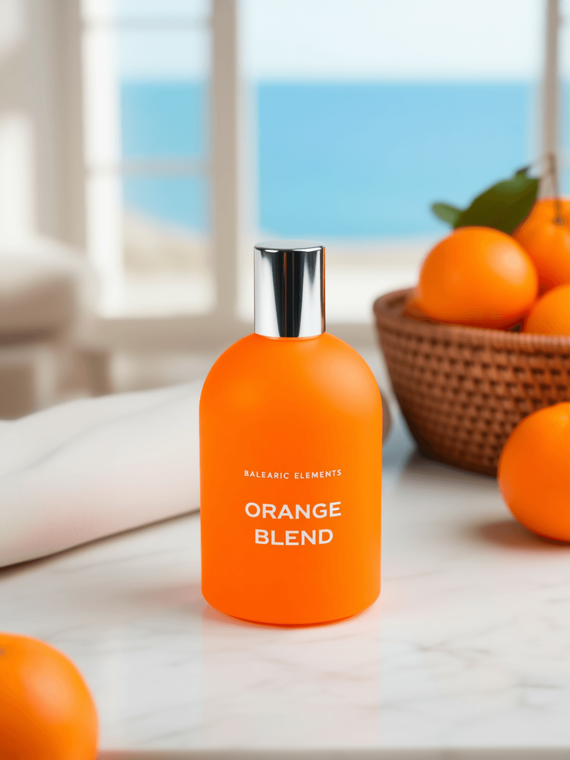 An orange bottle labeled 'BALEARIC ELEMENTS ORANGE BLEND' stands on a white marble surface with oranges and a woven basket in the background, near a bright window.
