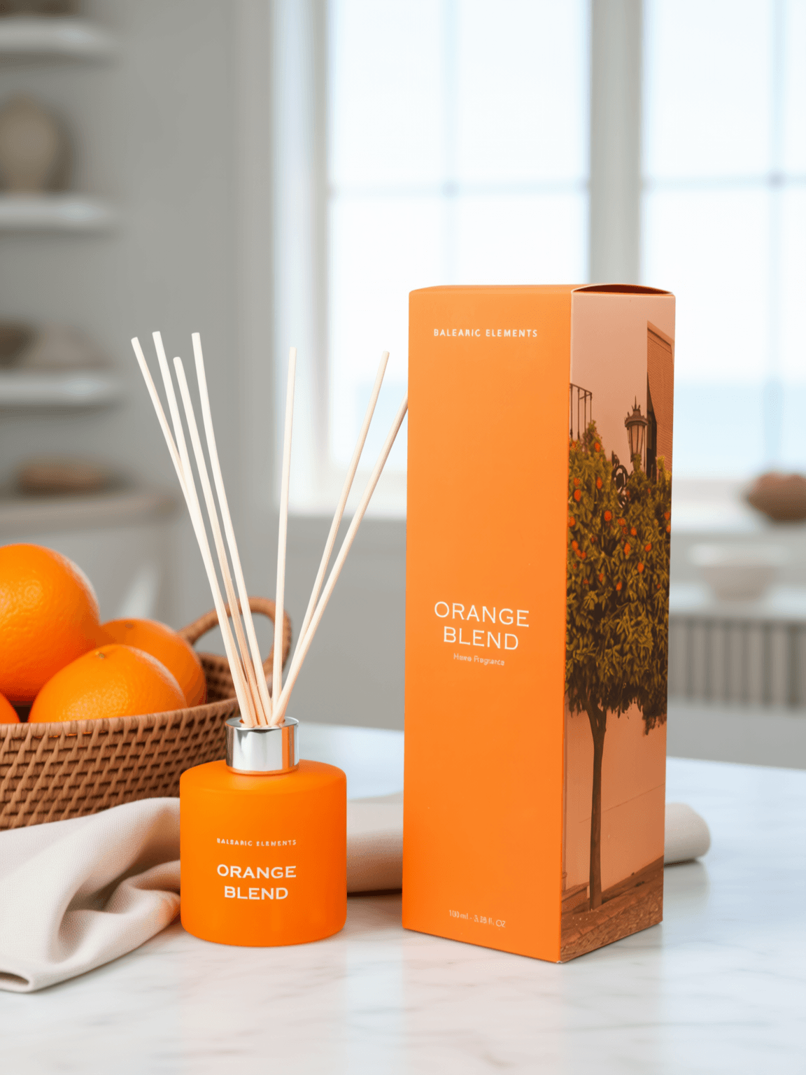 An orange reed diffuser bottle with wooden reeds stands beside its matching orange box on a light countertop. The packaging and bottle are labeled 'ORANGE BLEND' and 'BALEARIC ELEMENTS', with oranges and a basket visible in the softly blurred background.
