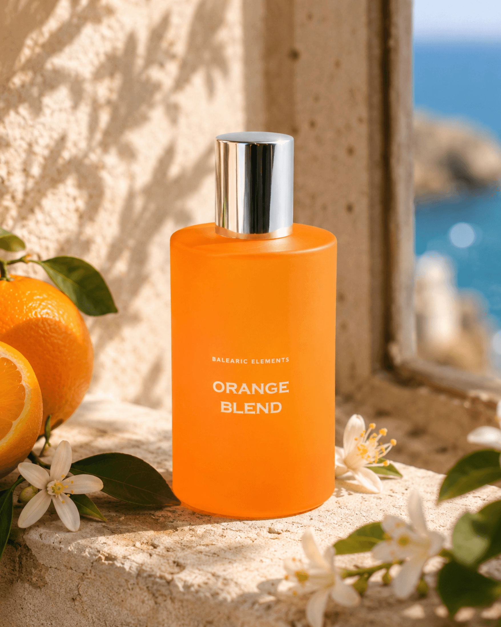 An orange bottle labeled 'BALEARIC ELEMENTS ORANGE BLEND' stands on a stone ledge outdoors, with oranges, white blossoms, and a blurred blue sea/window background.