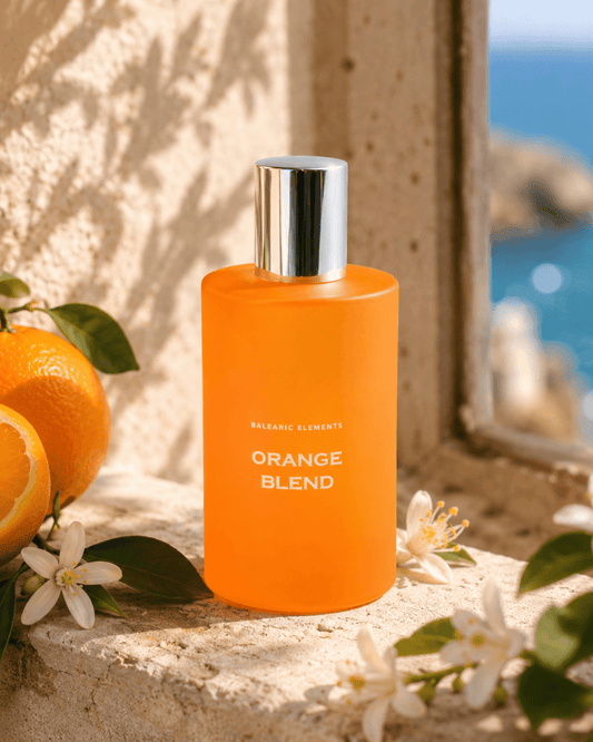 An orange bottle labeled 'BALEARIC ELEMENTS ORANGE BLEND' stands on a stone ledge outdoors, with oranges, white blossoms, and a blurred blue sea/window background.