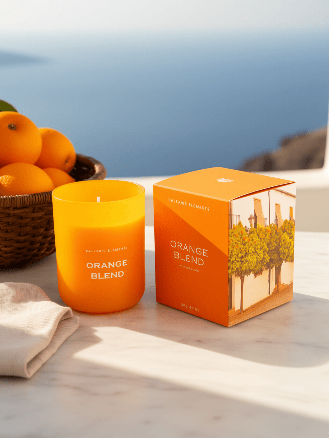 An orange glass candle labeled 'ORANGE BLEND' stands beside its matching orange box on a light marble surface, with oranges and a folded cloth nearby and a blurred ocean view in the background.