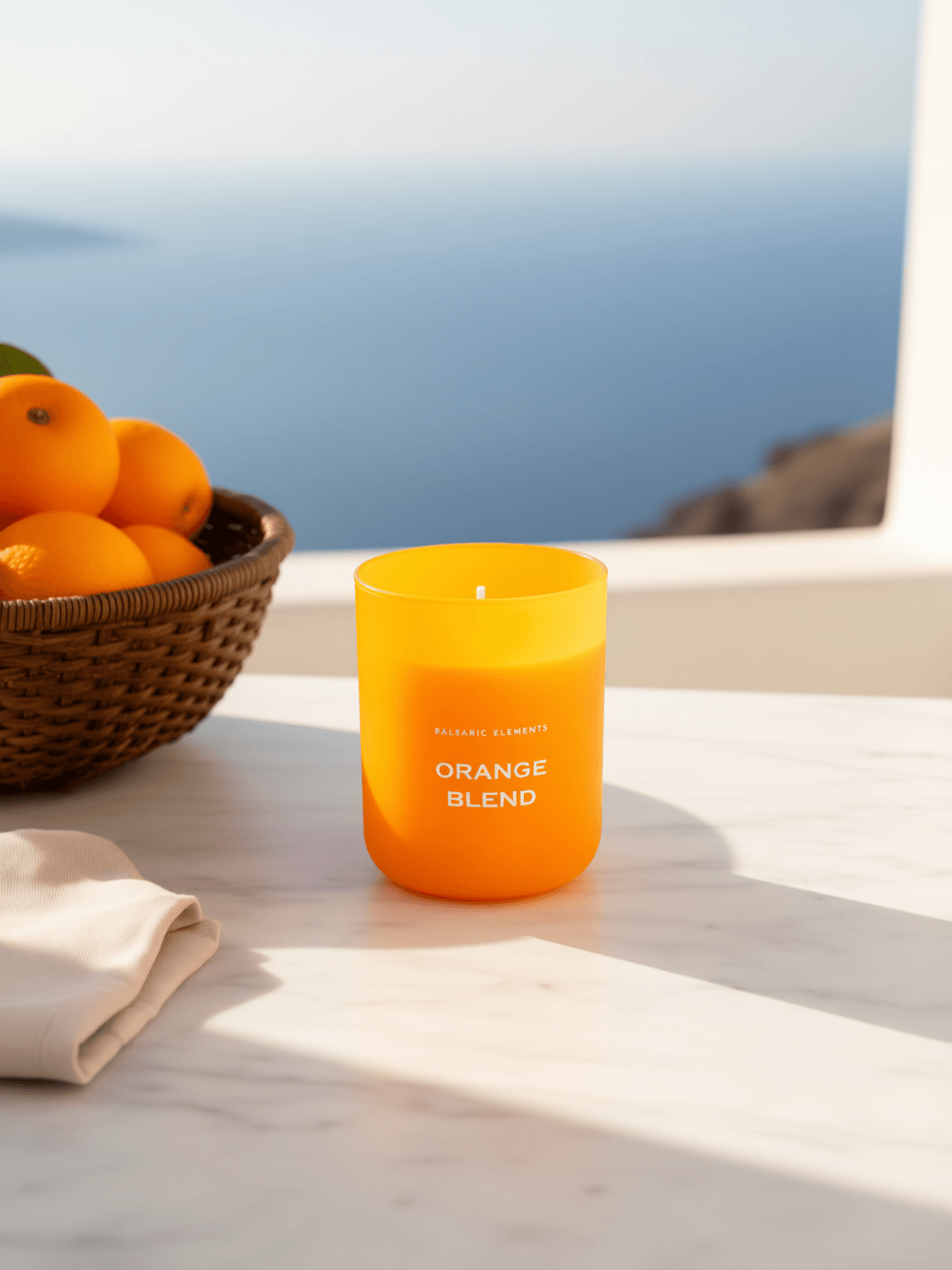 An orange-coloured scented candle labeled 'ORANGE BLEND' sits on a white marble surface with a basket of oranges and a folded cloth nearby, with a blurred blue sea background visible through a window.