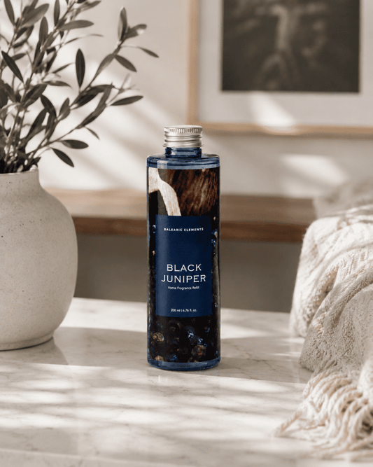 A bottle of Balearic Elements Black Juniper reed diffuser refill standing on a marble surface in a styled interior setting with a potted plant and a throw blanket.