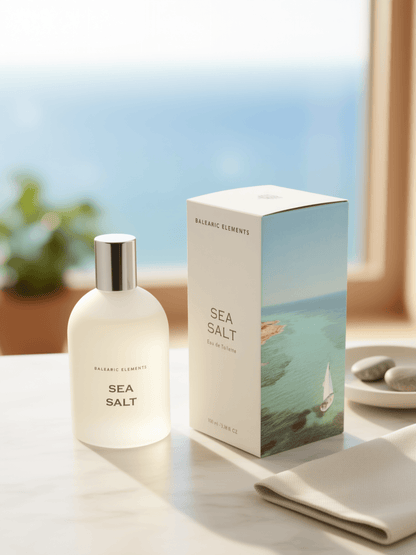 A frosted glass bottle labeled 'BALEARIC ELEMENTS SEA SALT' stands beside its matching product box on a light marble surface. The box features a coastal ocean illustration with a sailboat.