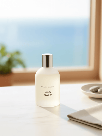 A frosted white bottle of Balearic Elements Sea Salt perfume with a silver cap, set on a light marble countertop with a blurred window and ocean view in the background.