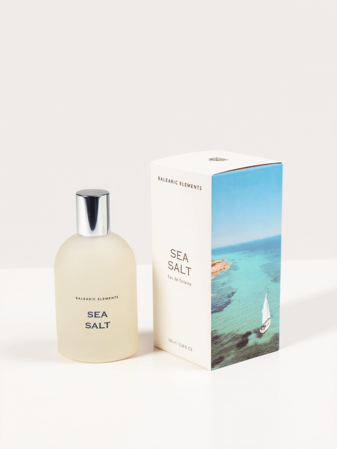 A frosted glass bottle of Balearic Elements Sea Salt Eau de Toilette next to its white packaging box featuring a blue ocean and sailboat illustration, on a clean white background.