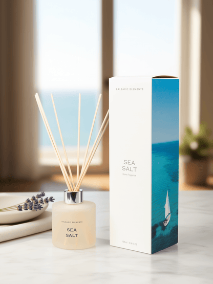 A reed diffuser bottle with pale beige liquid and wooden reeds stands beside its white and blue box on a light marble surface. The packaging features a blue ocean scene with a sailboat, and the diffuser is labeled 'BALEARIC ELEMENTS SEA SALT'.