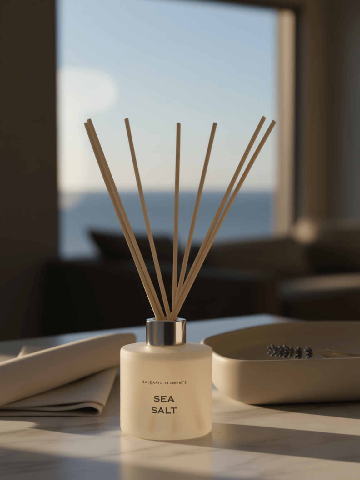 A sea salt reed diffuser bottle with natural wood reeds, placed on a table in a softly lit room with a blurred window and sofa in the background.