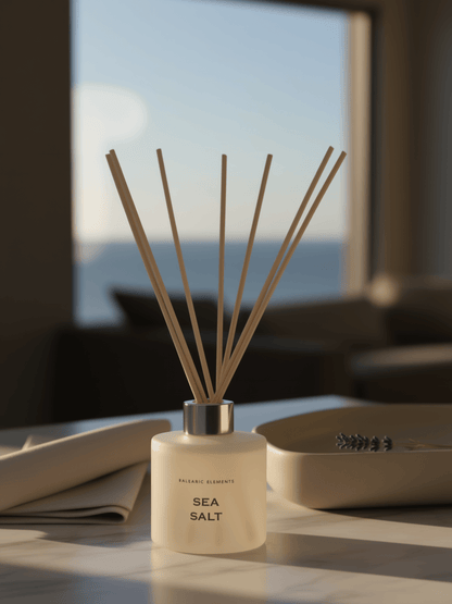 A sea salt reed diffuser bottle with natural wood reeds, placed on a table in a softly lit room with a blurred window and sofa in the background.