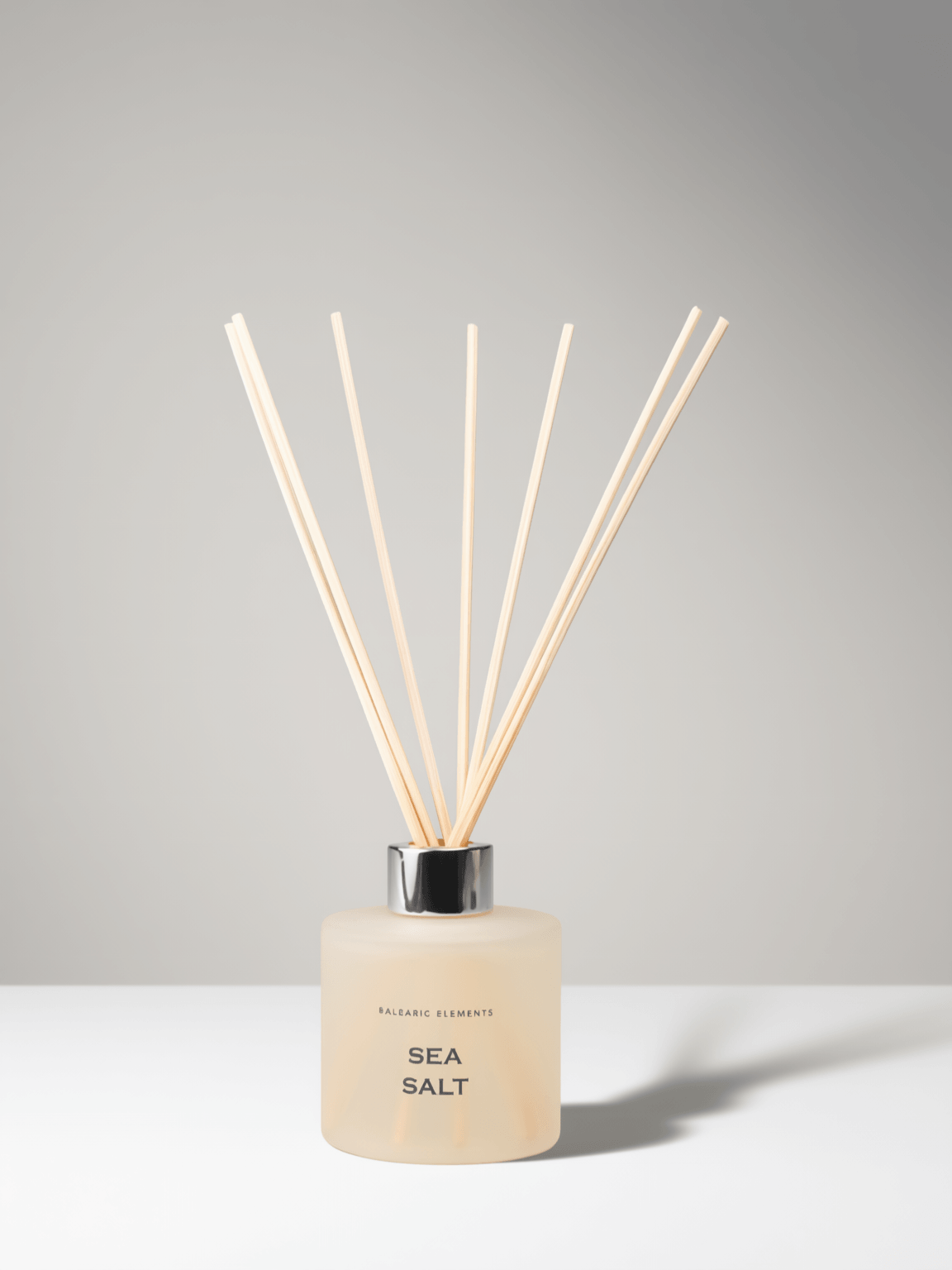 A reed diffuser bottle with a frosted pale beige glass body, black cap, and natural wood reeds, labeled 'SEA SALT' on a light gray background.