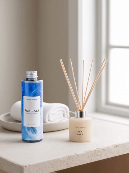 A bottle of Balearic Elements Sea Salt diffuser refill and a matching reed diffuser bottle with wooden reeds, styled on a light stone countertop with a folded white towel and dish in a softly lit room.