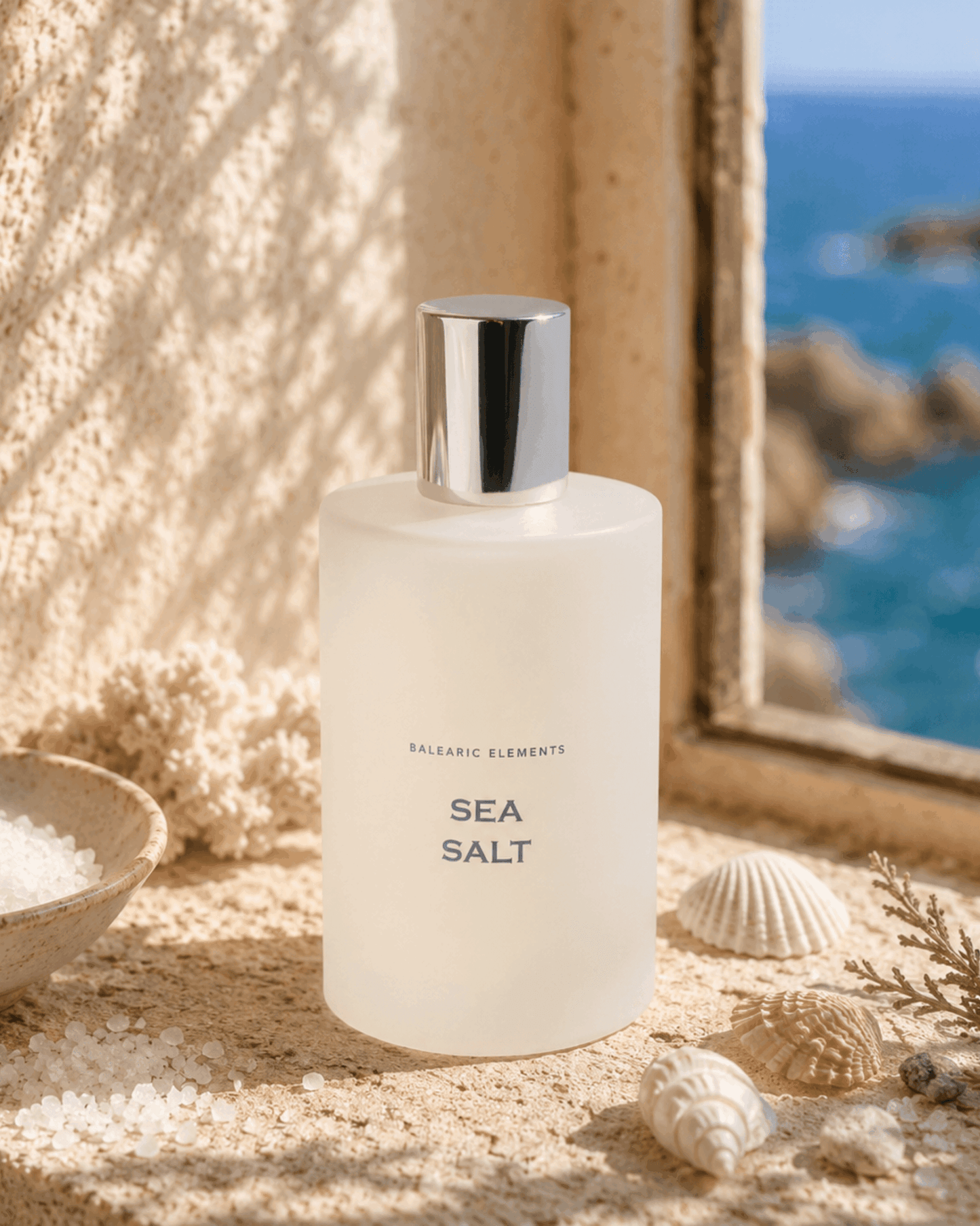 A frosted glass bottle labeled 'BALEARIC ELEMENTS SEA SALT' with a reflective silver cap, styled on a sandy surface with seashells and a bowl of salt, with a window view of the ocean in the background.