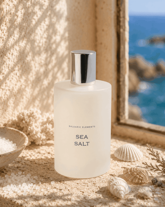 A frosted glass bottle labeled 'BALEARIC ELEMENTS SEA SALT' with a reflective silver cap, styled on a sandy surface with seashells and a bowl of salt, with a window view of the ocean in the background.