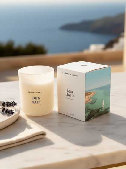 A frosted glass jar candle labeled 'SEA SALT' sits on a light stone surface beside its matching box, with a blurred coastal ocean scene in the background.