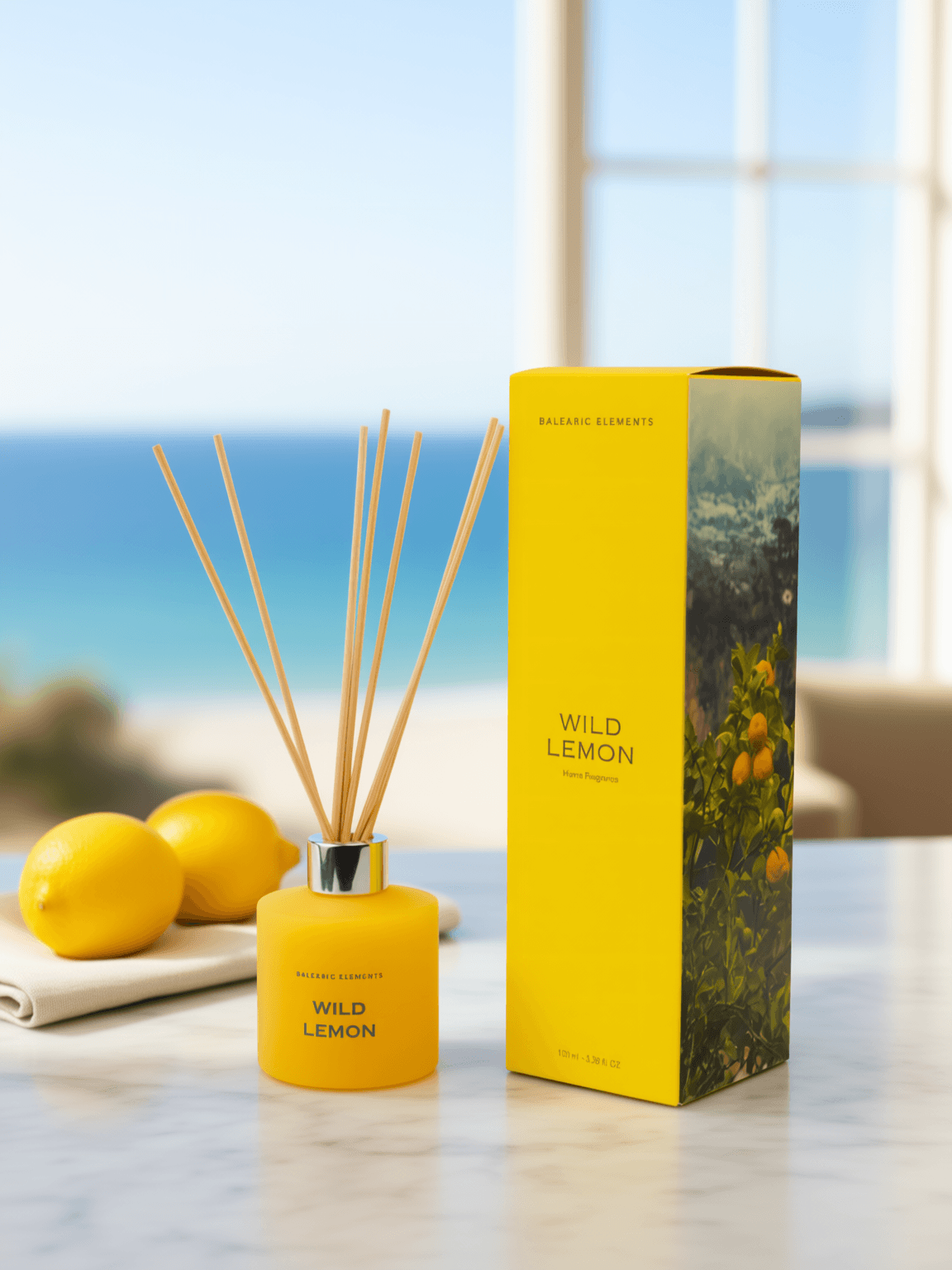 A yellow Balearic Elements Wild Lemon reed diffuser bottle with wooden reeds stands next to its matching yellow packaging box on a light marble surface, with lemons and a blurred ocean view in the background.