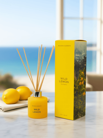 A yellow Balearic Elements Wild Lemon reed diffuser bottle with wooden reeds stands next to its matching yellow packaging box on a light marble surface, with lemons and a blurred ocean view in the background.