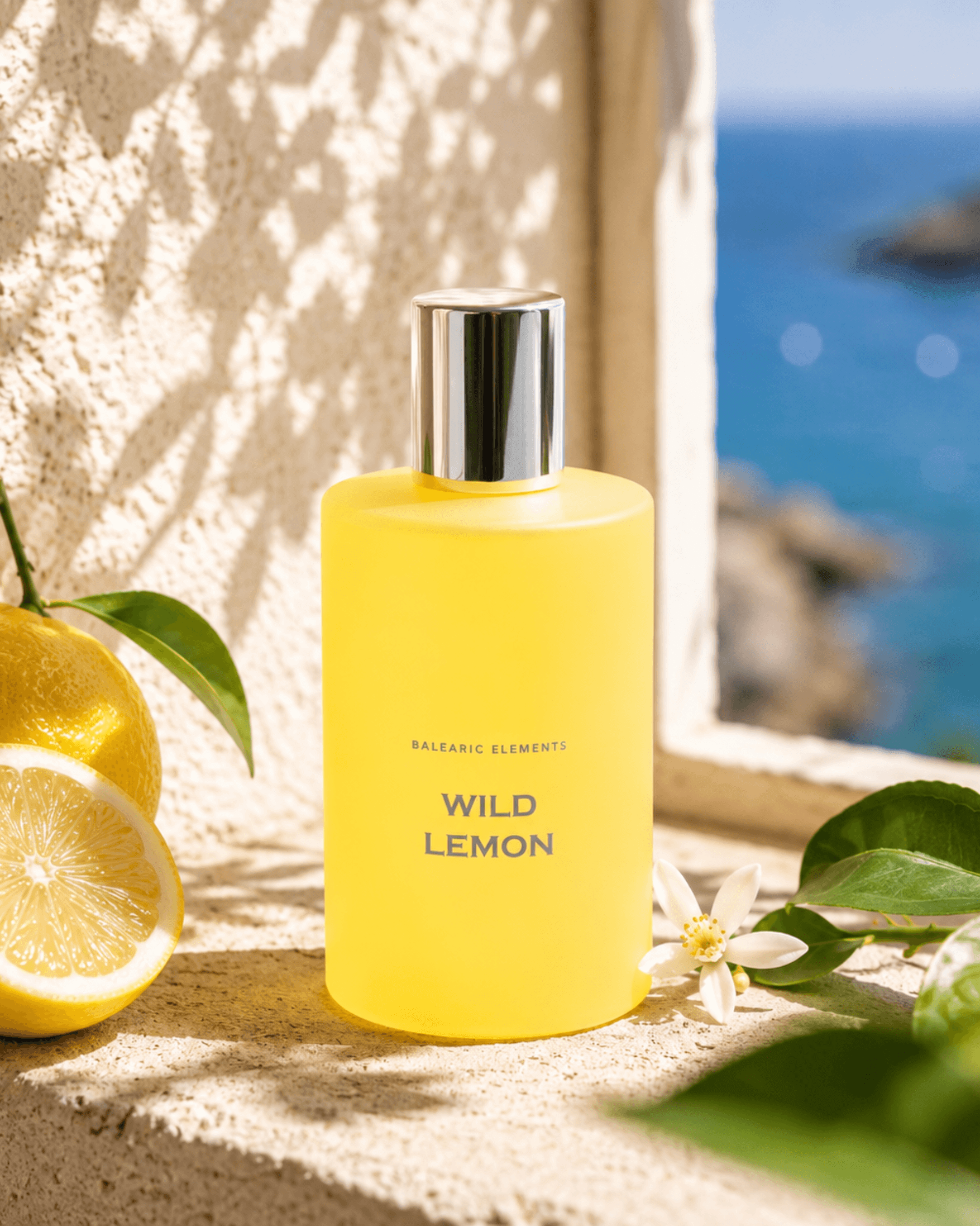 A frosted yellow bottle labeled 'WILD LEMON' by Balearic Elements stands on a stone ledge with lemons, green leaves, and a blue ocean background visible through a window.