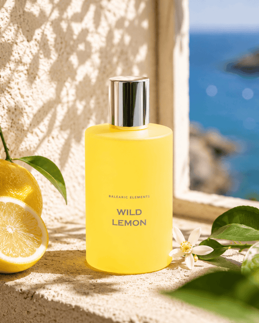 A frosted yellow bottle labeled 'WILD LEMON' by Balearic Elements stands on a stone ledge with lemons, green leaves, and a blue ocean background visible through a window.
