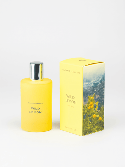 A yellow bottle of Wild Lemon Room Spray by Balearic Elements stands next to its matching yellow packaging box featuring a lemon and mountain landscape illustration, set against a plain white background.