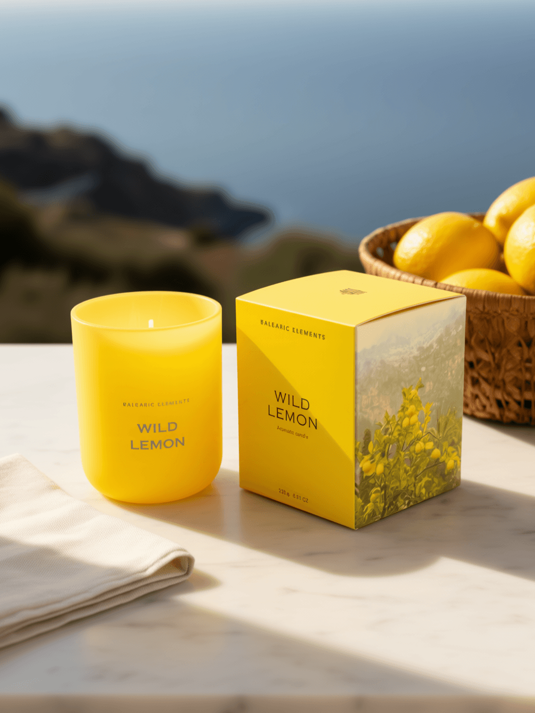 A bright yellow scented candle and its matching yellow box labeled 'WILD LEMON' sit on a light stone surface outdoors, with a basket of lemons and a blurred coastal background.