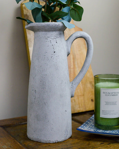 Grey rustic pitcher holding faux flowers on a wooden surface next to a candle