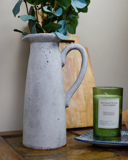 Grey rustic pitcher holding faux flowers on a wooden surface next to a candle with wooden grazing board in the background