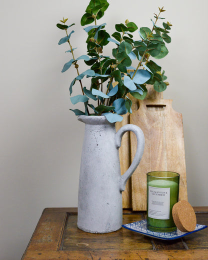 Gray pitcher with green leaves, green candle, and wooden grazing board on a wooden table.