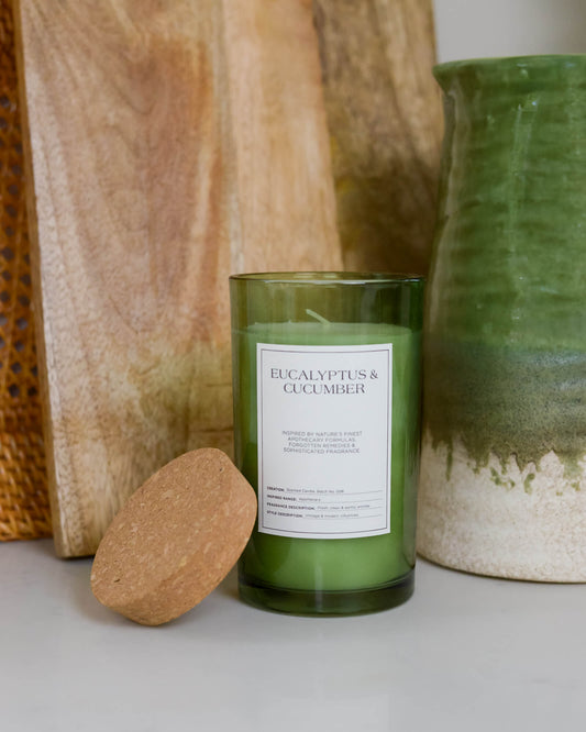 Glass jar candle with cork lid labelled 'Eucalyptus & Cucumber" on a marble kitchen worktop with grazing board and green vase 