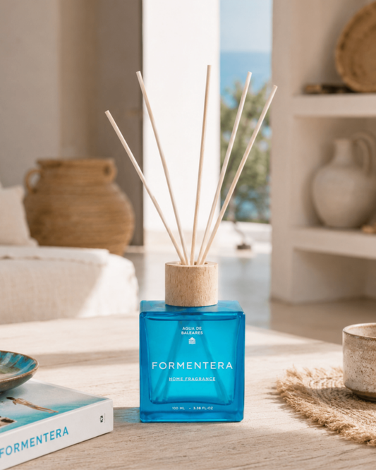 A Formentera reed diffuser with a bright blue glass bottle and natural wood reeds, styled on a light wooden table in a bright, modern interior setting.