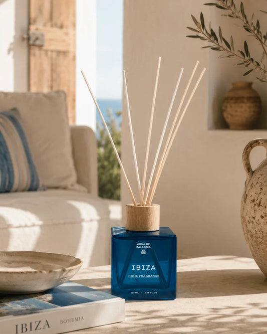 A blue reed diffuser bottle labeled IBIZA with several natural reed sticks, placed on a tabletop in a sunlit interior setting with a sofa, bowl, and decorative vase in the background.