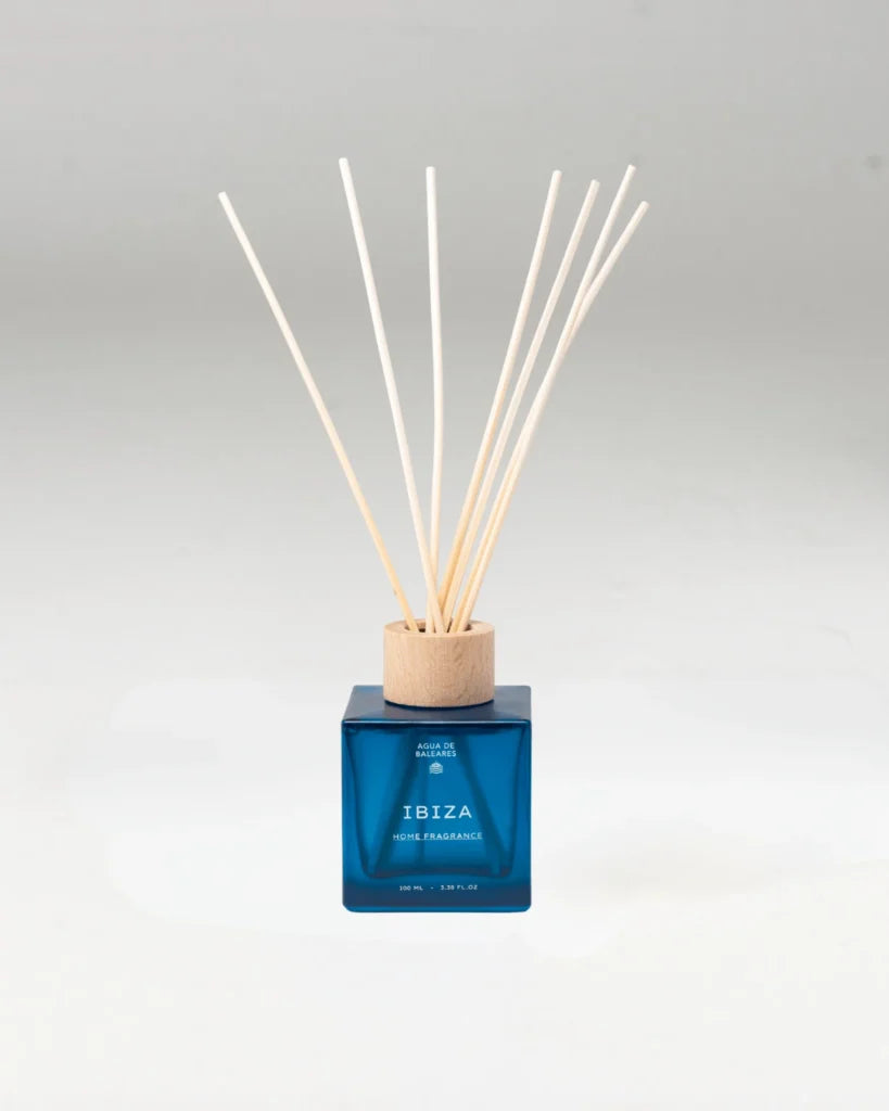 A reed diffuser bottle with a translucent blue glass body, a light wood cap, and multiple natural reed sticks, centered on a light gray background.