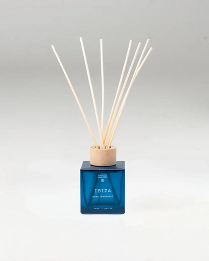 A reed diffuser bottle with a translucent blue glass body, a light wood cap, and multiple natural reed sticks, centered on a light gray background.