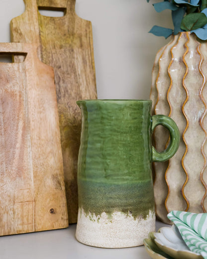 Green ceramic pitcher with wooden cutting boards and a decorative vase in the background.