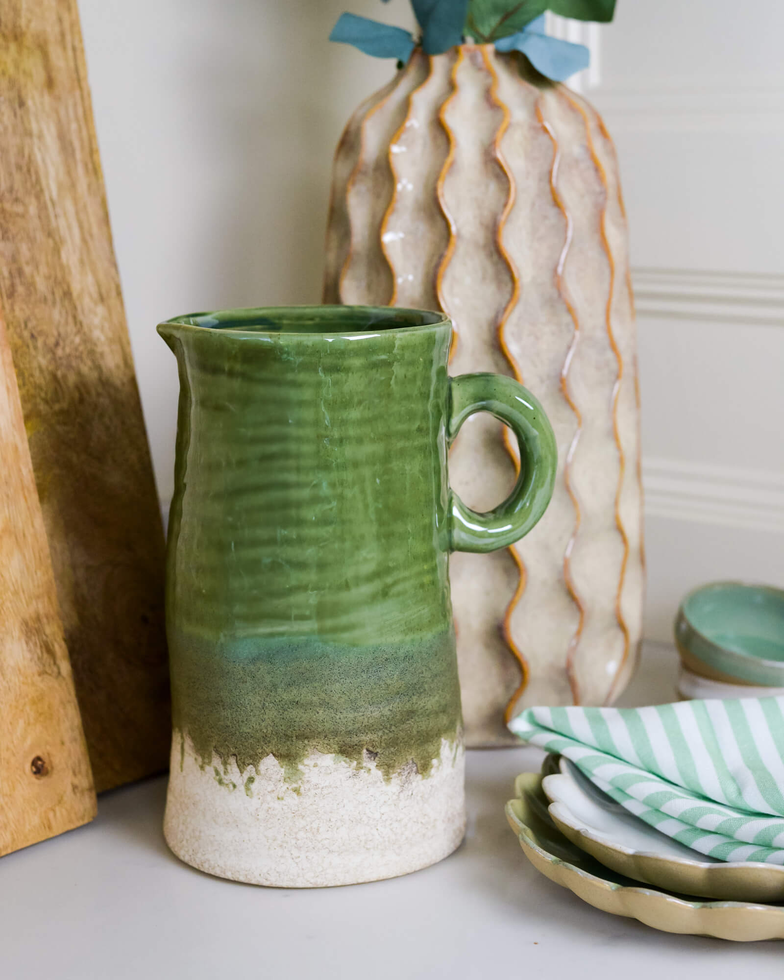 Large olive green ceramic jug with a textured surface on a table with a vase and plates in the background.