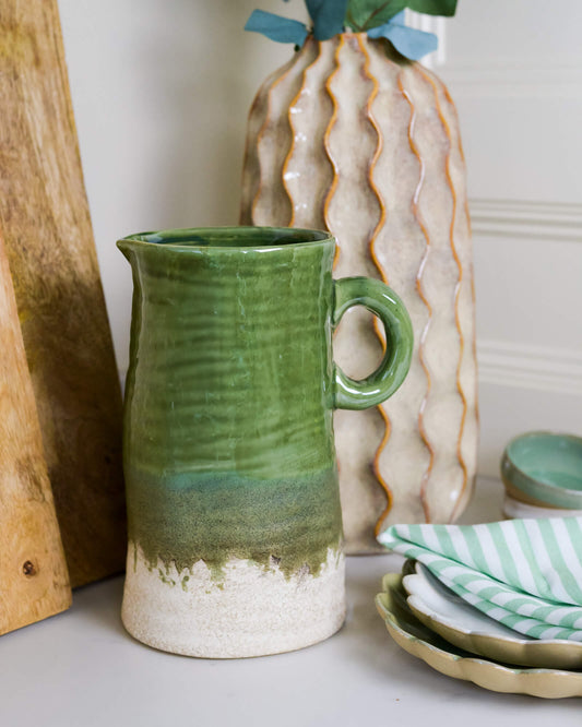 Large olive green ceramic jug with a textured surface on a table with a vase and plates in the background.