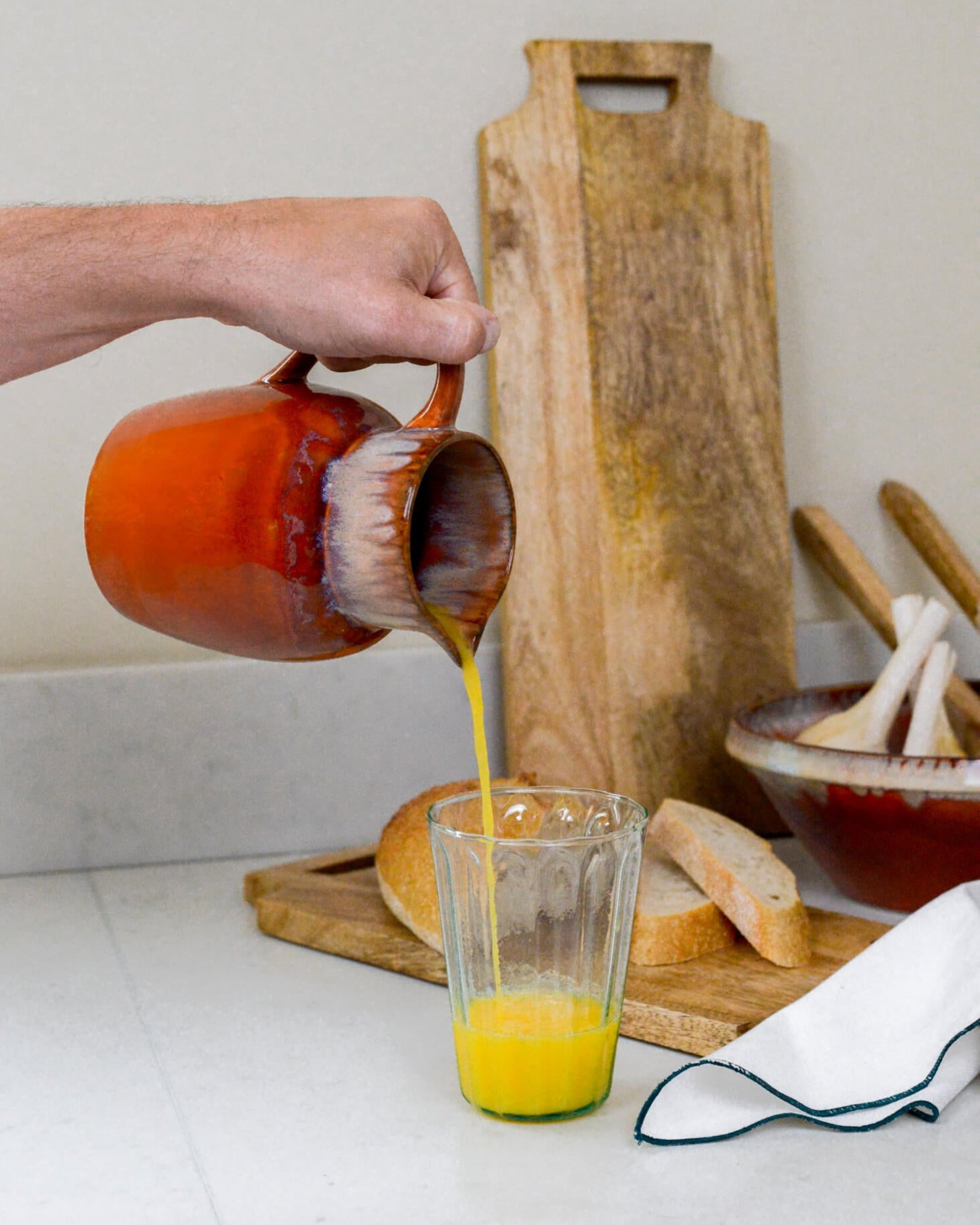 Person pouring orange juice from a ceramic pitcher into a glass on a kitchen counter.