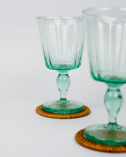 Two green wine glass goblets on woven coasters with a white background