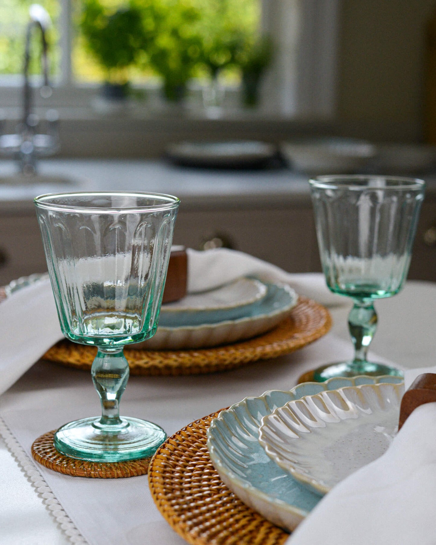 Two green glass goblet wineglasses on a table with ceramic plates and a blurred kitchen background