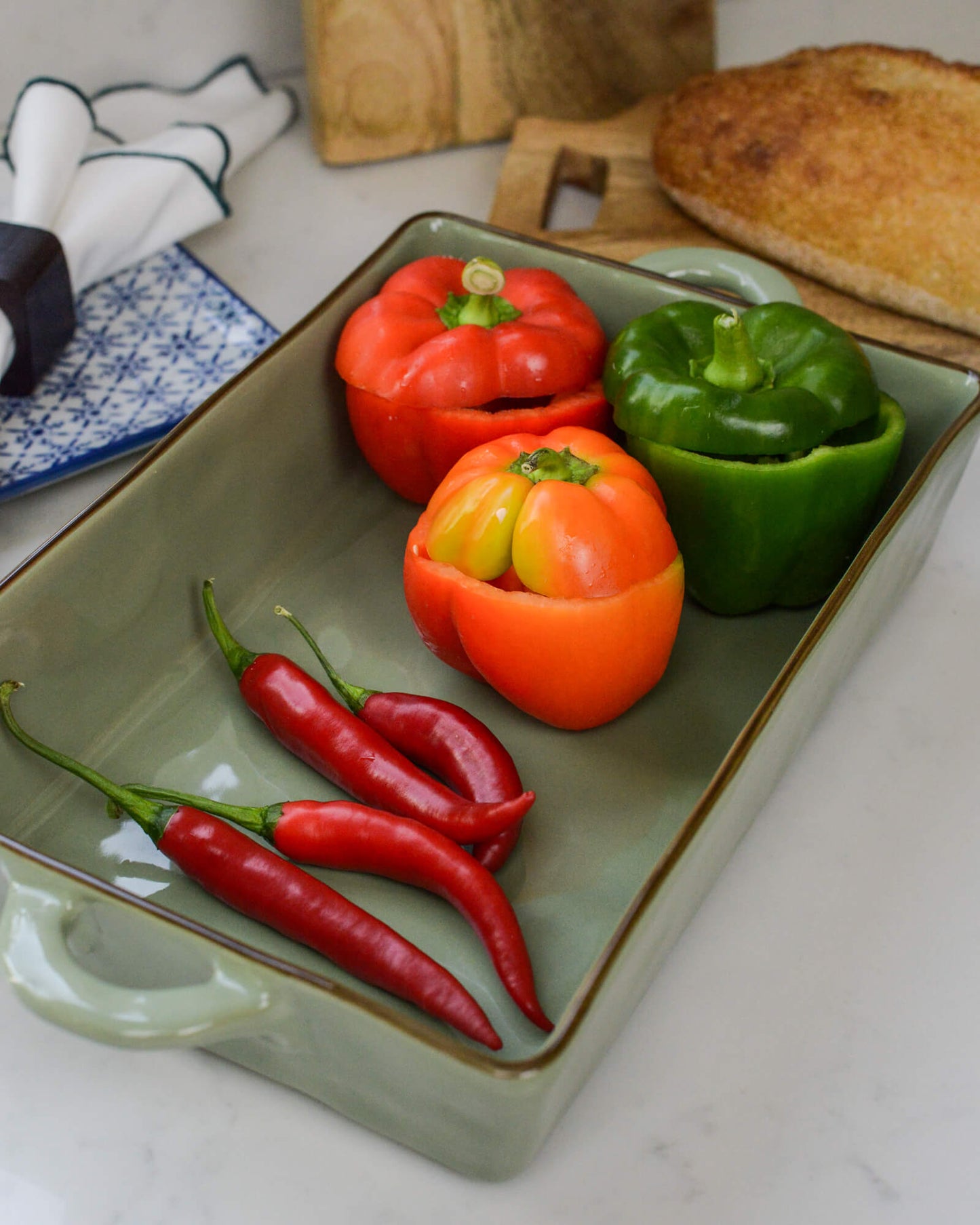 Stuffed bell peppers and red chilies in an olive green ceramic baking dish with bread in the background.