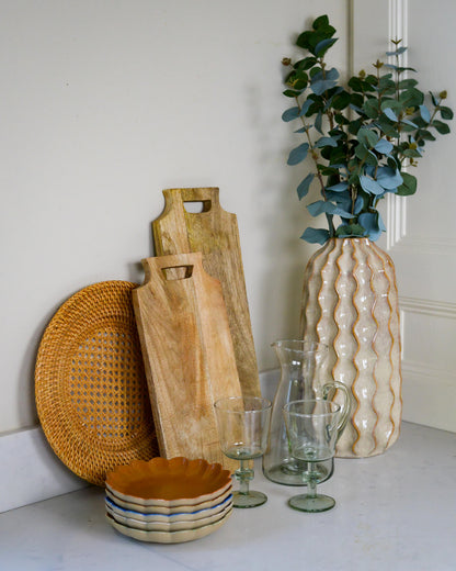 Decorative items including wooden cutting boards, a rattan charger plate, a vase with greenery, and glassware on a white marble surface.