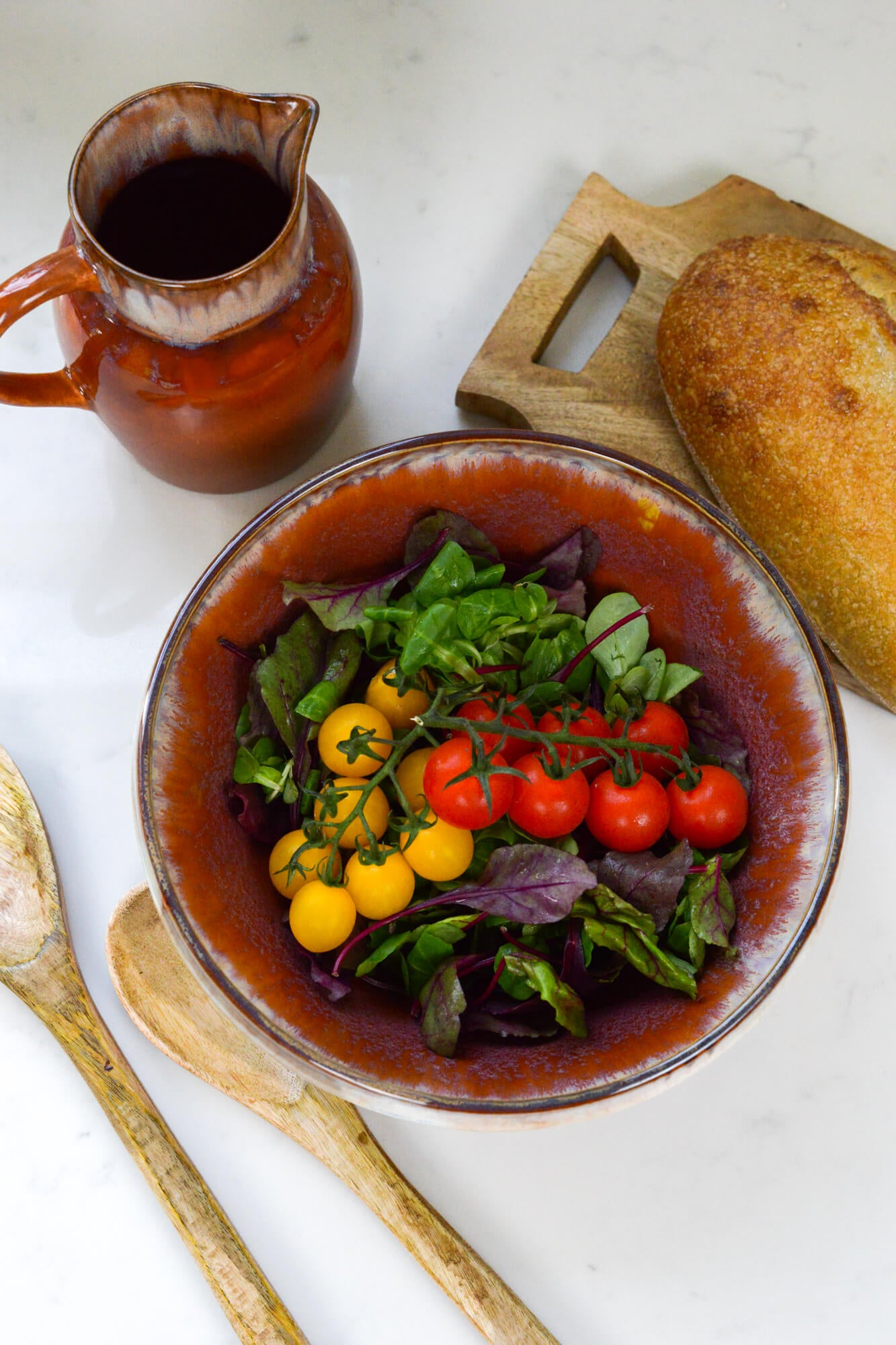 Salad bowl with, bread, and a pitcher on a white surface with salad servers 