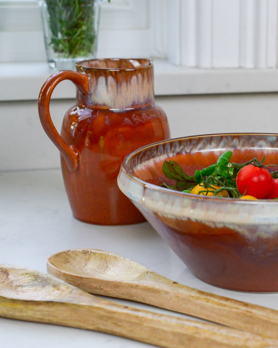 Ceramic pitcher and bowl with tomato-based dish on a white surface with wooden salad servers